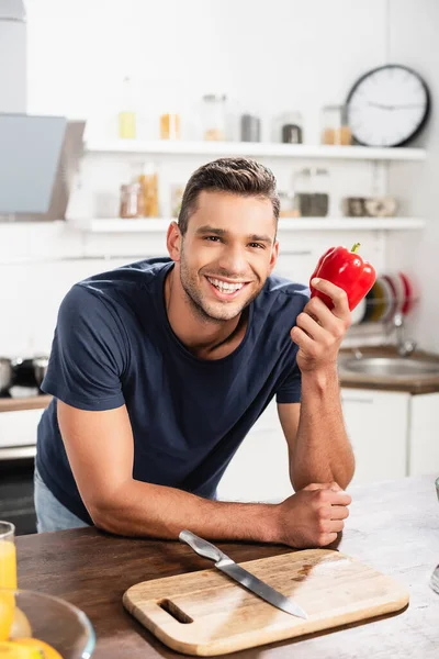 Uomo sorridente che tiene la paprica vicino al tagliere e coltello sul tavolo della cucina — Foto stock