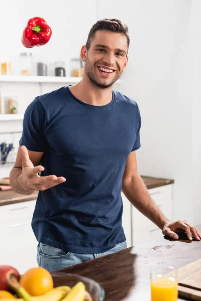 Homem sorridente jogando pimentão perto de um copo de suco de laranja e frutas em primeiro plano turvo — Fotografia de Stock