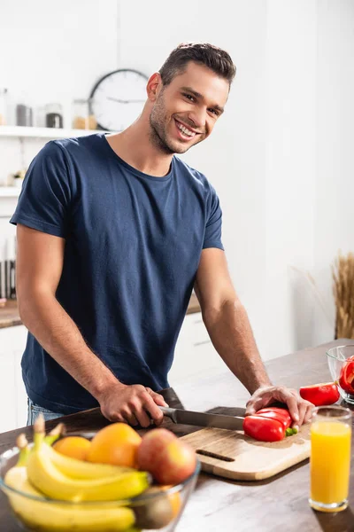 Smiling man cutting paprika near fruits and glass of orange juice on blurred foreground - foto de stock