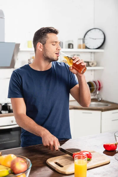 Homme buvant de la bière en coupant du poivron près du jus d'orange et des fruits au premier plan flou — Photo de stock