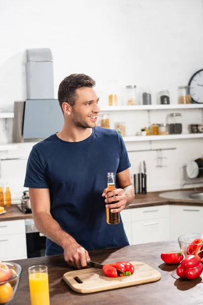 Un homme souriant tenant une bouteille de bière tout en coupant du poivron près du jus d'orange et des fruits au premier plan flou — Photo de stock