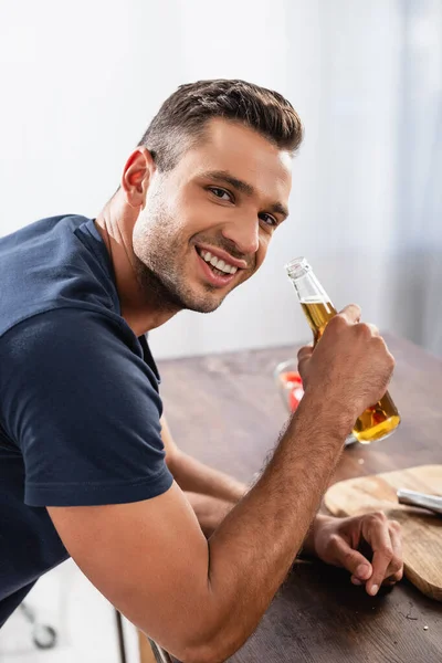 Uomo sorridente in possesso di bottiglia di birra vicino tagliere su sfondo sfocato in cucina — Foto stock