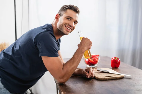 Vue latérale d'un homme souriant tenant une bouteille de bière près de la planche à découper et du poivron sur fond flou — Photo de stock