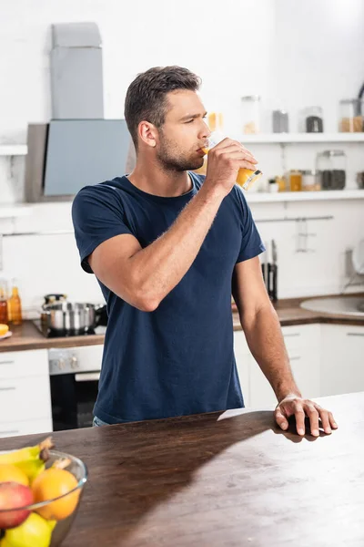 Young man drinking orange juice near fresh fruits on blurred foreground on table — Stock Photo