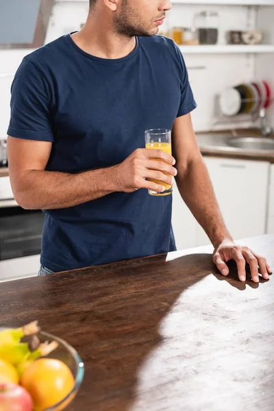 Vista cortada do homem segurando vidro de suco de laranja perto de frutas em primeiro plano desfocado na cozinha — Fotografia de Stock