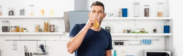 Junger Mann schaut in die Kamera, während er Orangensaft in der Küche trinkt, Banner — Stockfoto