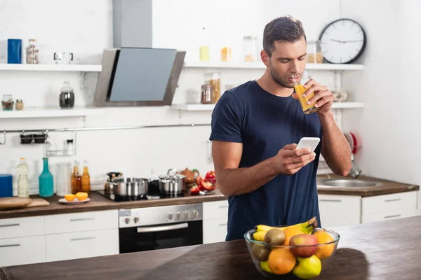 Jeune homme utilisant un smartphone tout en buvant du jus d'orange près de fruits au premier plan flou — Photo de stock