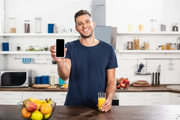 Homme souriant montrant smartphone avec écran blanc près du verre de jus d'orange et de fruits dans la cuisine — Photo de stock