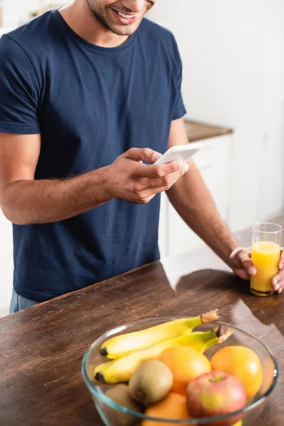 Vista cortada do homem sorridente usando smartphone perto de vidro de suco de laranja e frutas frescas em primeiro plano desfocado — Fotografia de Stock