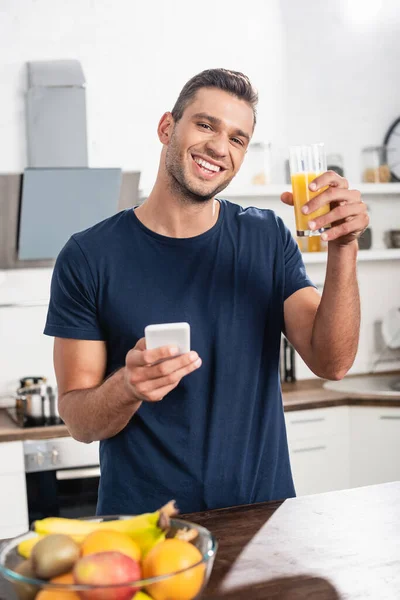 Uomo sorridente che tiene il bicchiere di succo d'arancia e smartphone vicino ai frutti maturi su primo piano sfocato — Foto stock