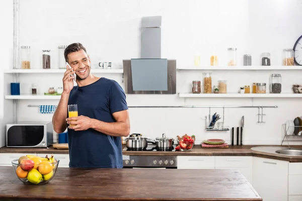 Smiling man talking on smartphone while holding glass of orange juice near fruits in kitchen — Stock Photo