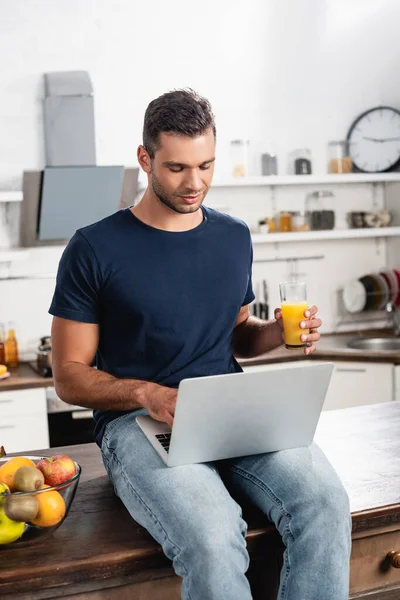 Man with laptop and orange juice sitting near fruits on kitchen table — Stock Photo