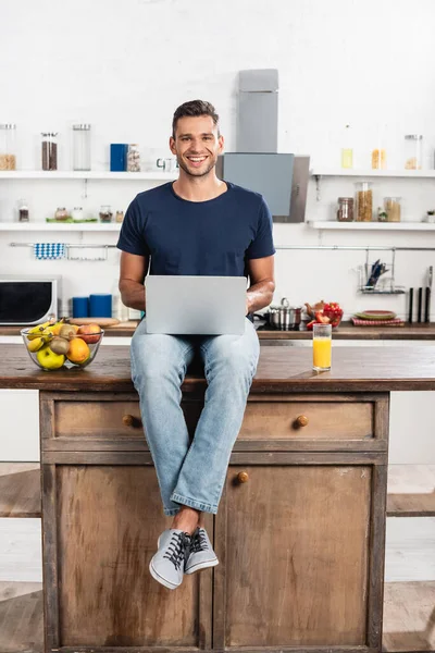 Uomo sorridente guardando la fotocamera durante l'utilizzo di laptop vicino succo d'arancia e frutta in cucina — Foto stock