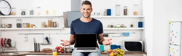 Smiling man pointing with hands near laptop with blank screen, fruits and orange juice in kitchen, banner - foto de stock