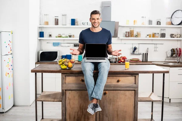 Smiling man pointing with hands near laptop with blank screen, orange juice and ripe fruits in kitchen — Stock Photo