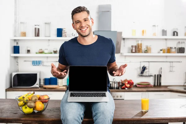 Homem alegre apontando com as mãos perto do laptop com tela em branco, frutas e suco de laranja na mesa da cozinha — Fotografia de Stock