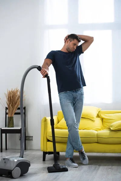 Tired man standing near vacuum cleaner in living room - foto de stock