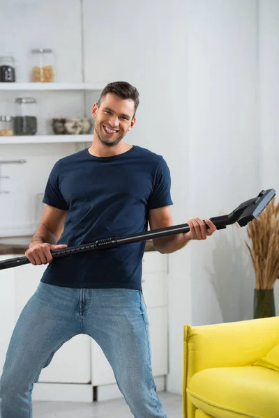 Cheerful man holding brush of vacuum cleaner near couch at home - foto de stock
