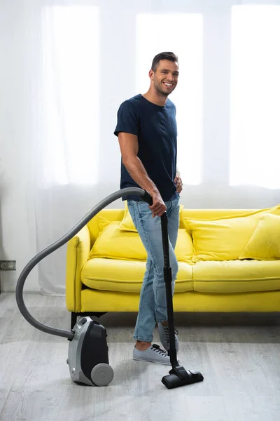 Lächelnder Mann mit Hand auf Hüftbürste des Staubsaugers zu Hause — Stockfoto