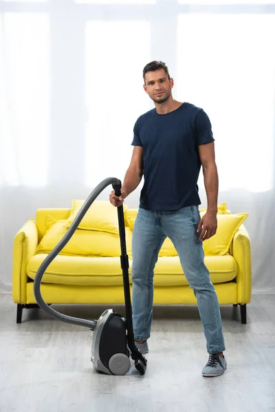 Young man holding vacuum cleaner near sofa in living room — Stock Photo