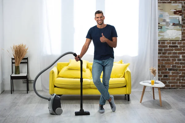 Smiling man showing thumb up while standing near vacuum cleaner in living room - foto de stock