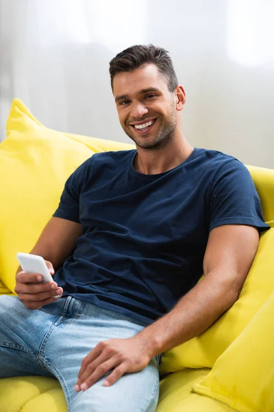 Uomo sorridente che usa lo smartphone seduto sul divano giallo — Foto stock
