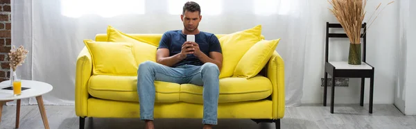 Jeune homme assis sur le canapé tout en utilisant un smartphone près du verre de jus d'orange sur la table basse, bannière — Photo de stock