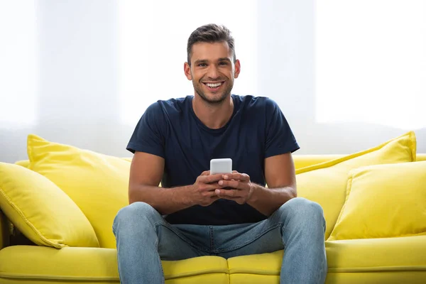 Jeune homme souriant à la caméra tout en utilisant un smartphone sur le canapé — Photo de stock