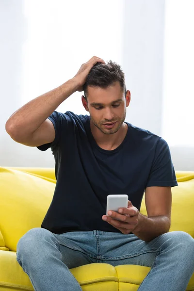 Man with hand near head using smartphone on yellow sofa — Stock Photo
