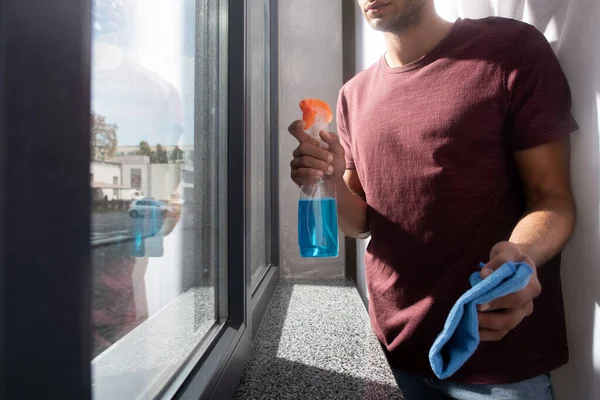 Cropped view of man holding detergent and rag near window on blurred foreground - foto de stock