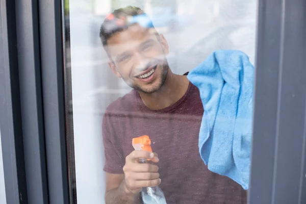 Young man smiling at camera while cleaning window with rag and detergent — Stock Photo