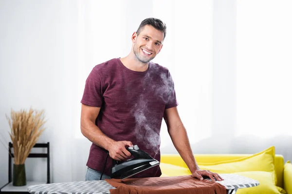 Uomo sorridente che tiene il ferro con il vapore vicino ai vestiti a bordo a casa — Foto stock
