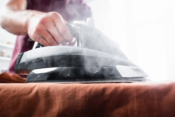 Cropped view of iron with steam on clothes in hand of man on blurred background — Stock Photo