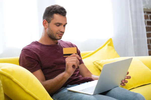 Jeune homme tenant une carte de crédit tout en utilisant un ordinateur portable sur le premier plan flou — Photo de stock