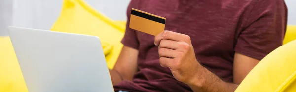 Vue recadrée de l'homme tenant la carte de crédit près de l'ordinateur portable sur le canapé sur fond flou, bannière — Photo de stock