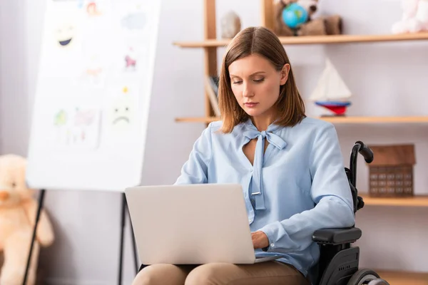 Psicologa femminile che utilizza il computer portatile mentre seduto in sedia a rotelle con ufficio offuscato sullo sfondo — Foto stock