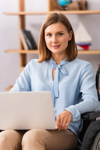 Позитивная женщина-психолог с ноутбуком, смотрящая в камеру, сидя в инвалидной коляске на размытом фоне — стоковое фото