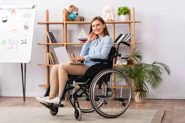Full length of smiling psychologist with laptop looking at camera while sitting in wheelchair in office — Stock Photo