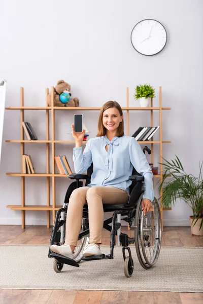 Longitud completa de una psicóloga sonriente que muestra un teléfono inteligente con pantalla en blanco mientras está sentada en silla de ruedas en la oficina - foto de stock