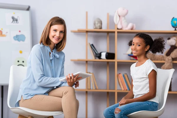 Lächelnder Psychologe mit digitalem Tablet und afrikanisch-amerikanisches Mädchen, das während einer Beratung auf verschwommenem Hintergrund in die Kamera blickt — Stockfoto