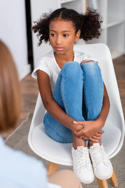 Chica afroamericana seria abrazando las piernas mientras está sentado en la silla durante la consulta con el psicólogo borroso en primer plano - foto de stock
