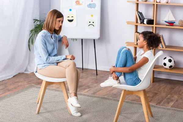 Psicólogo mirando a las niñas afroamericanas abrazando las piernas mientras se sienta en la silla durante la consulta en la oficina - foto de stock