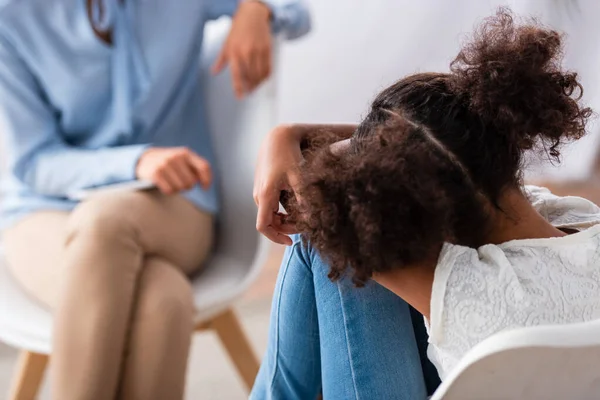 Visão traseira da menina afro-americana deprimida com psicólogo desfocado em segundo plano — Fotografia de Stock
