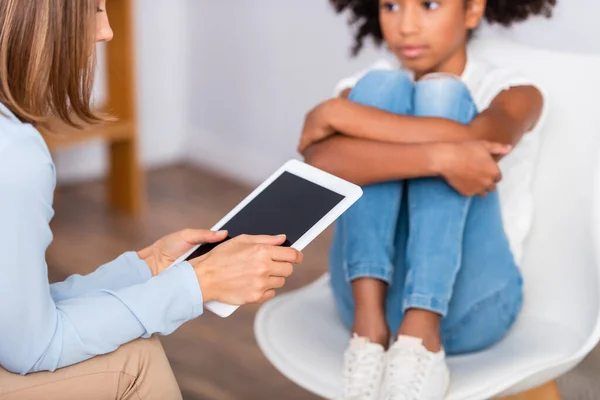 Vista cortada do psicólogo segurando tablet digital enquanto sentado perto de menina americana africana abraçando as pernas no fundo borrado — Fotografia de Stock
