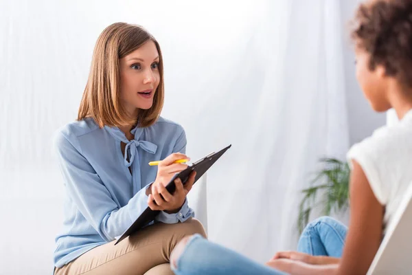 Psychologue surpris écrivant sur presse-papiers tout en regardant la fille afro-américaine pendant la consultation sur le premier plan flou — Photo de stock