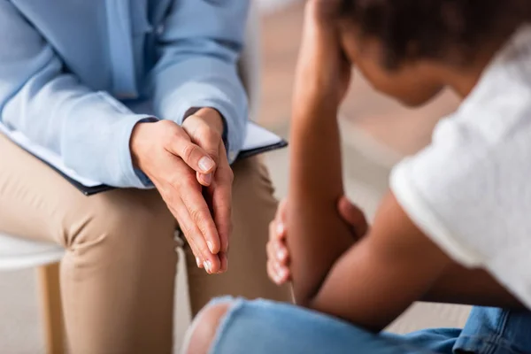 Vue recadrée du psychologue avec des mains priantes assis pendant la consultation avec une fille afro-américaine floue au premier plan — Photo de stock
