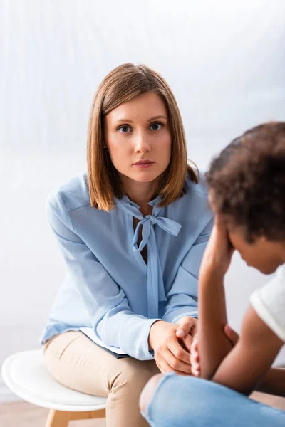 Psychologin blickt in die Kamera, während sie auf einem Stuhl neben verschwommenem afrikanisch-amerikanischem Mädchen sitzt, das im Vordergrund weint — Stockfoto