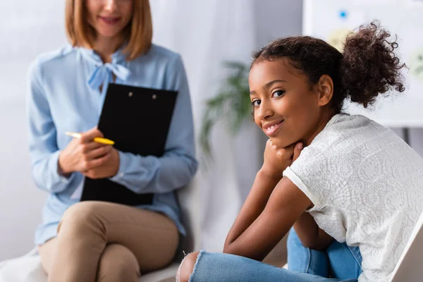 Happy african american girl looking at camera during consultation with blurred psychologist on background — Stock Photo
