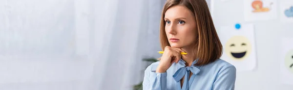 Psicóloga mujer reflexiva sosteniendo la pluma y mirando hacia otro lado con oficina borrosa en el fondo, pancarta - foto de stock