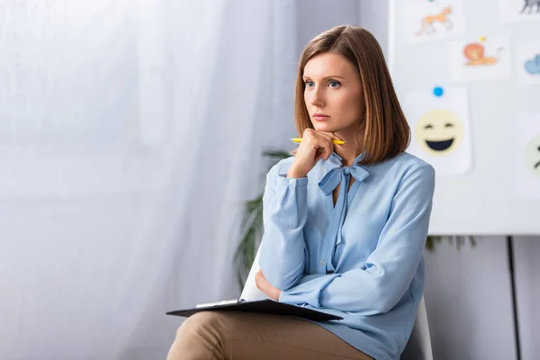 Psicóloga mujer pensativa con lápiz y portapapeles mirando hacia otro lado mientras está sentada en la silla con la oficina borrosa en el fondo - foto de stock